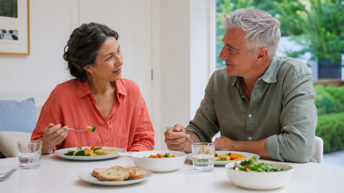 Two older adults in coral and sage shirts sharing dinner and conversation at a white table beside a garden window.
